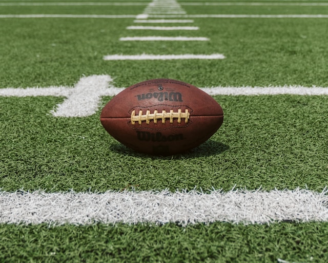 Close-up of a brown Wilson football on green turf at the 1-yard line with white field markings.