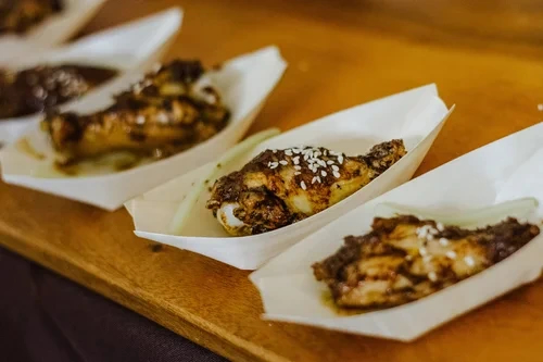 Chicken wings seasoned and served in disposable paper trays on a wooden surface at Death by Chocolate event