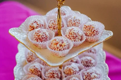 Two-tier tray with white and pink coconut-covered dessert balls in paper cups on a purple cloth.