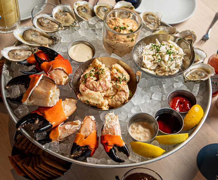 Seafood platter with crab claws, oysters on ice, dipping sauces, lemon wedges, and seafood salads in bowls.