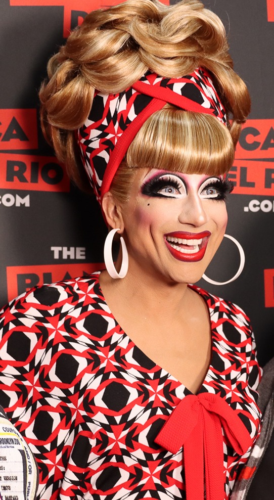 Drag queen with vibrant makeup, a voluminous blonde hairdo, red and black patterned outfit, and large white earrings.