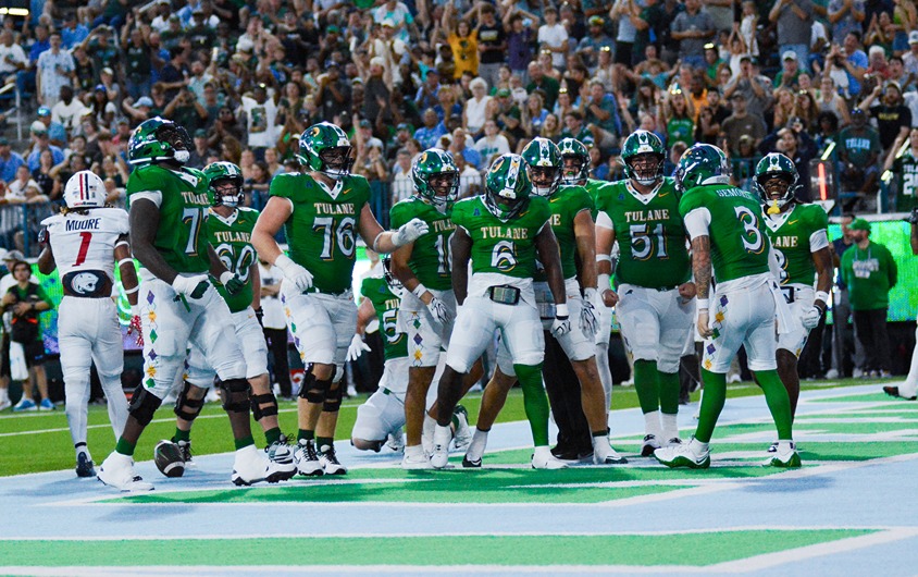 Tulane football players in green uniforms celebrate a touchdown in the end zone with a cheering crowd behind them.