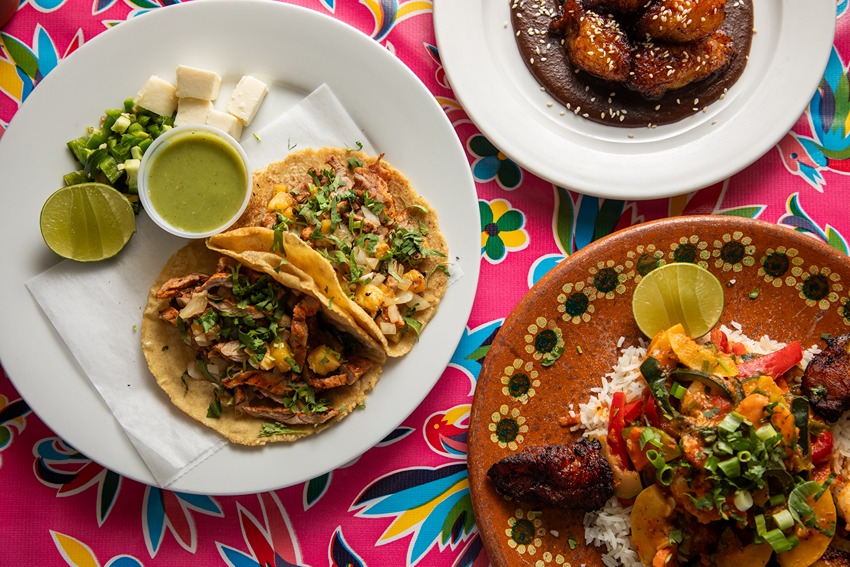 Two tacos with meat and pineapple, green sauce, cheese cubes, rice with vegetables, plantains on colorful tablecloth