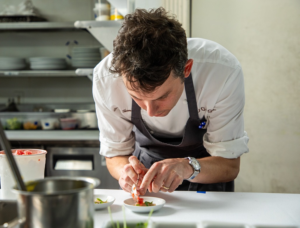 Chef in white shirt and black apron carefully plating food in a professional kitchen setting.