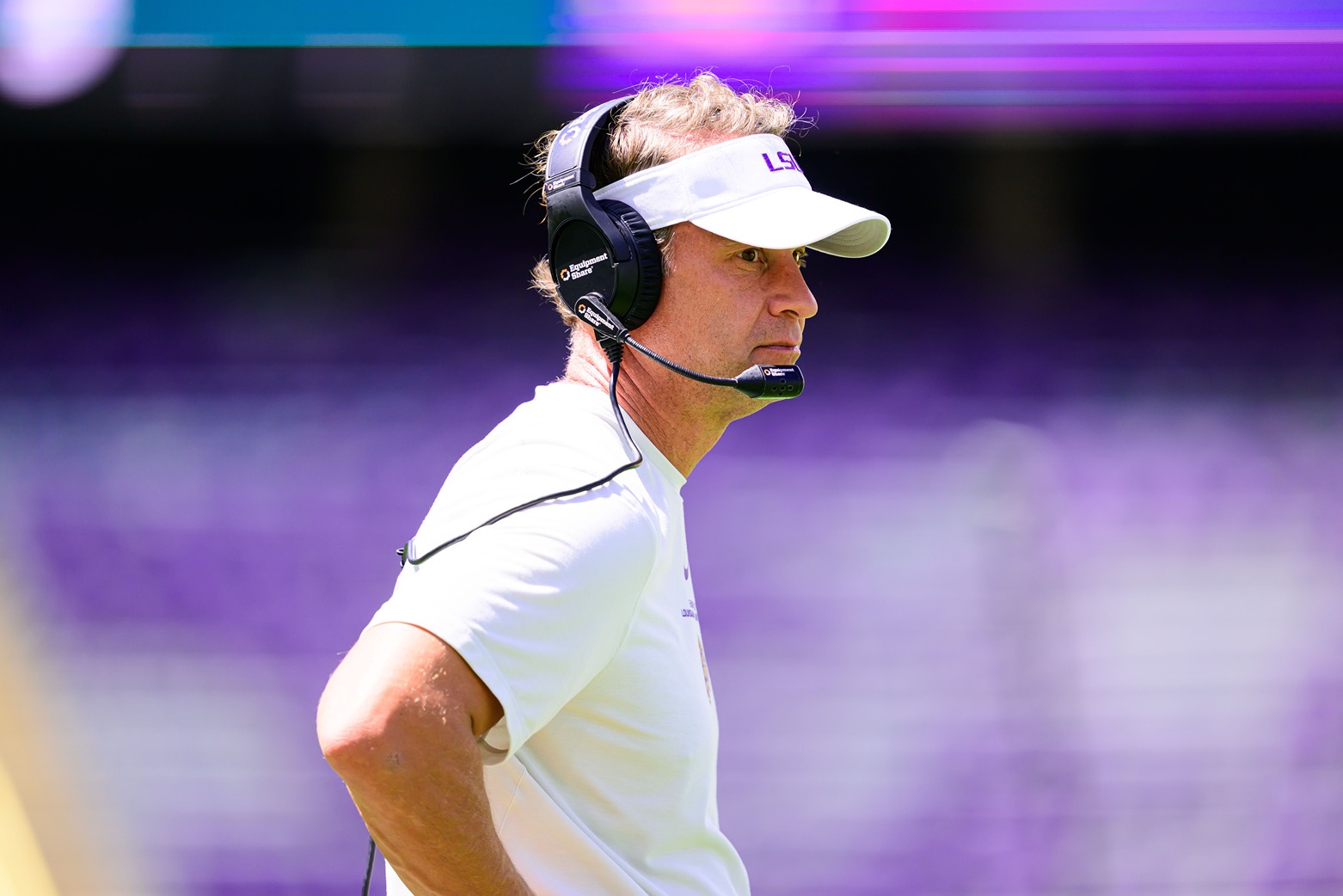 Football coach wearing LSU visor and headset stands on field focused during game day.
