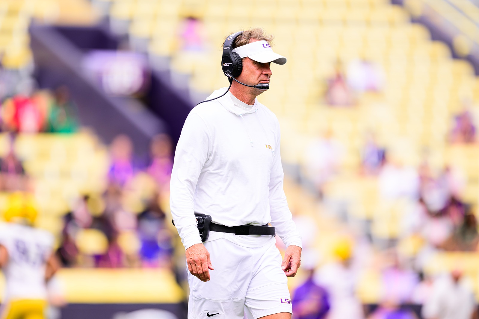 LSU football coach in white outfit and visor wearing headset during game on sidelines with blurred stadium background