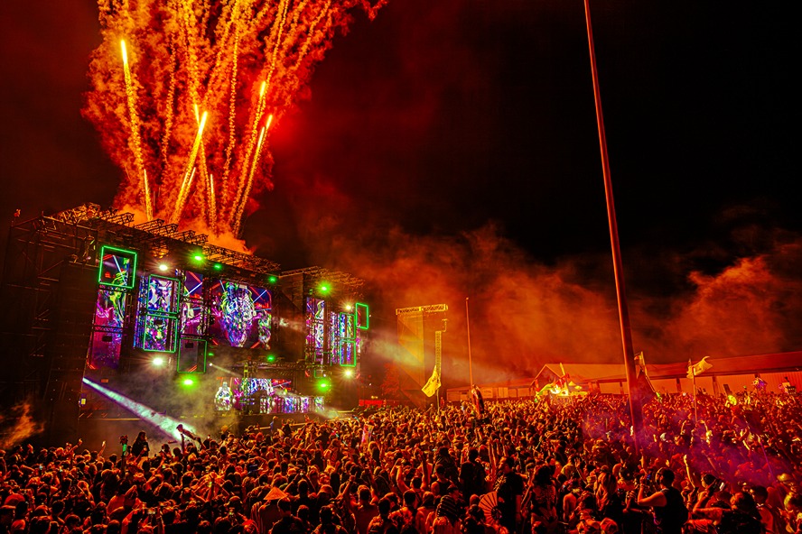 Crowded outdoor music festival stage with colorful lights, large audience, and fireworks lighting up the night sky