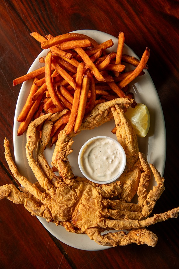 Plate of crispy fried soft shell crab with sweet potato fries, dipping sauce, and a lemon wedge on wooden table.