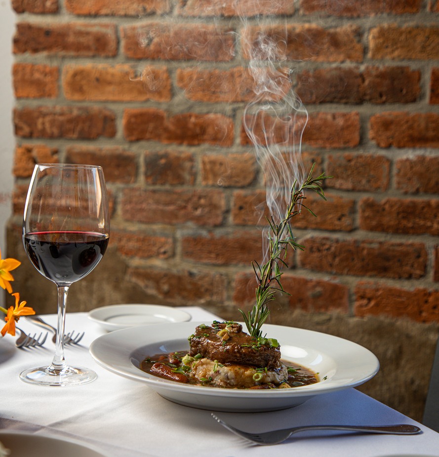 Plate of gourmet steak with steaming rosemary garnish and glass of red wine on white tablecloth by brick wall