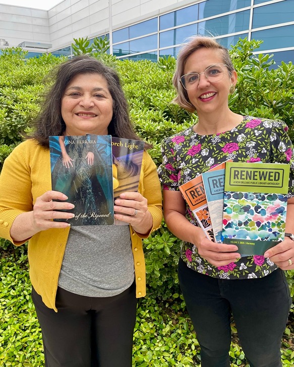 Two women smiling and holding various books and anthologies outdoors with green foliage background.