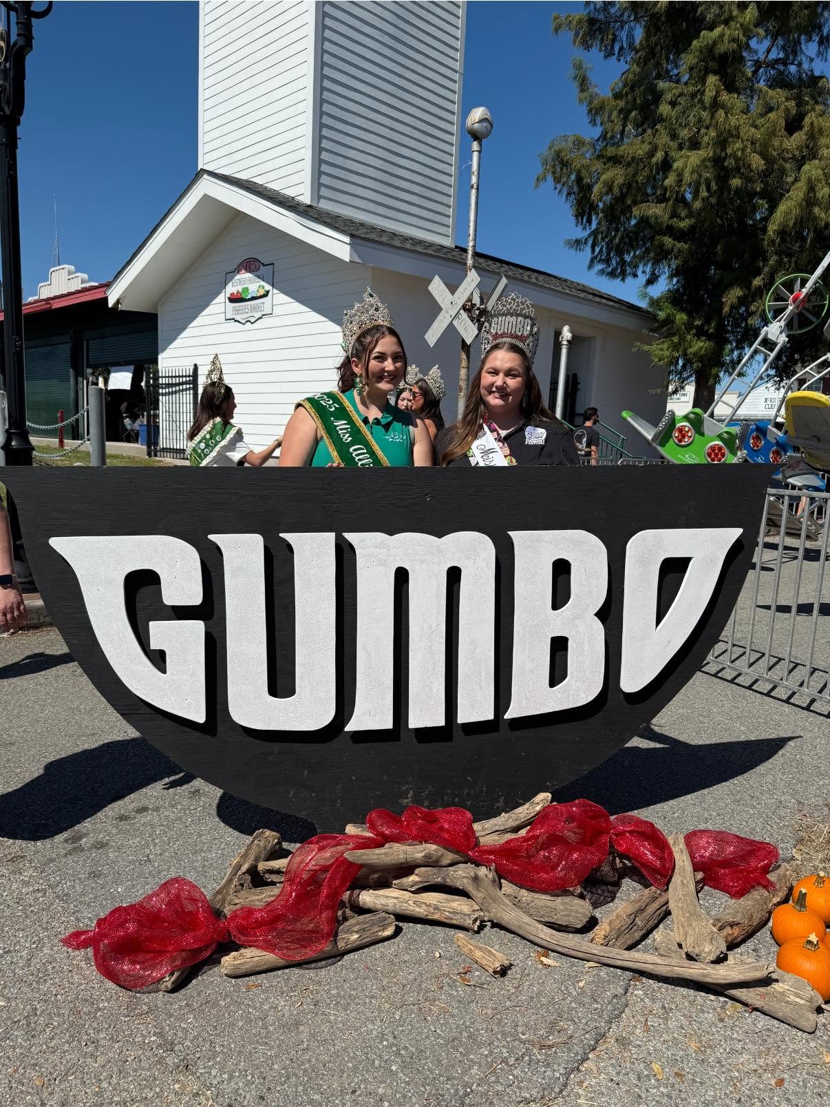 Two women wearing crowns and sashes stand behind a large black gumbo sign decorated with red mesh and wood on a sunny day.