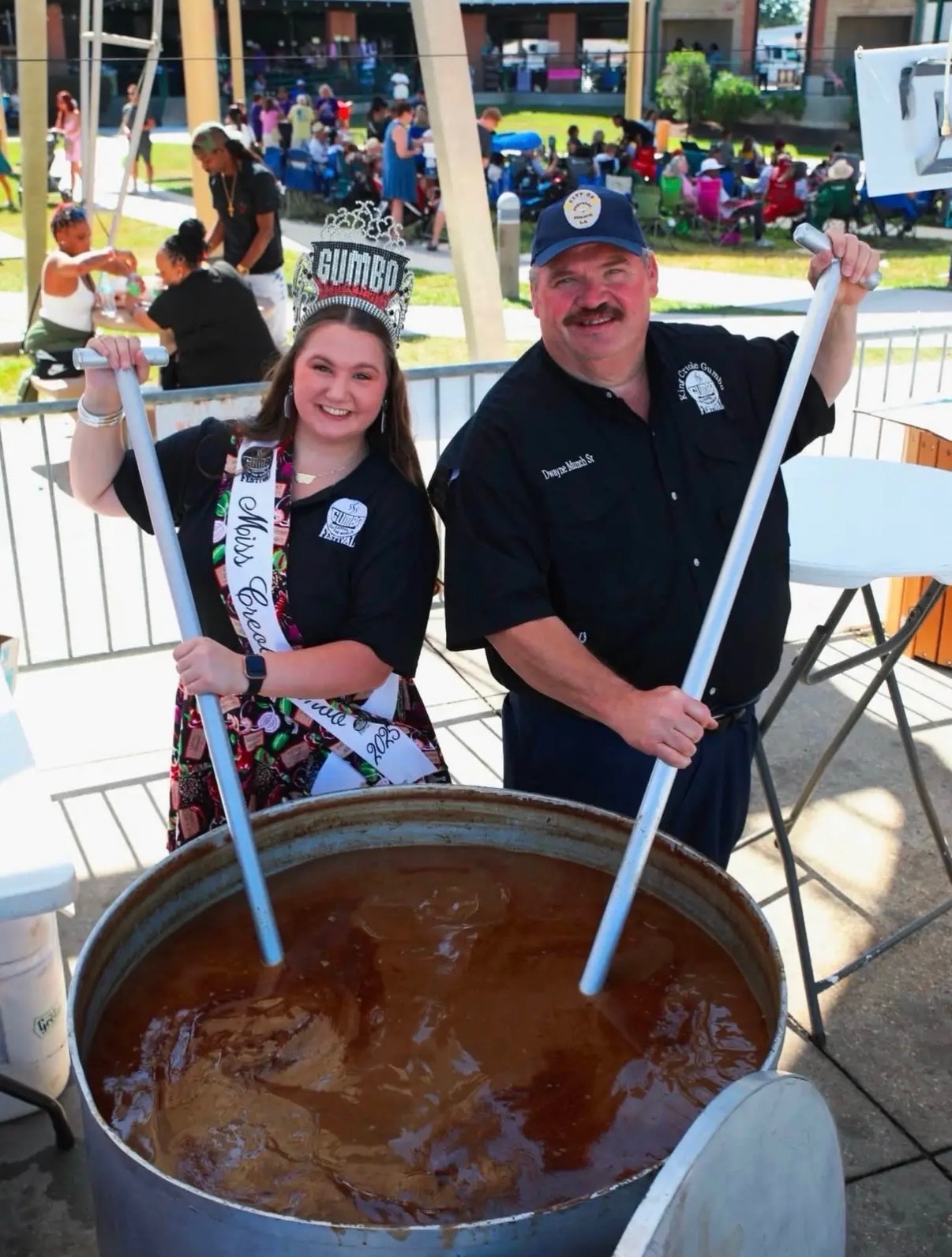 Miss Crawfish and a man stir a large pot of gumbo at an outdoor festival with people in the background.