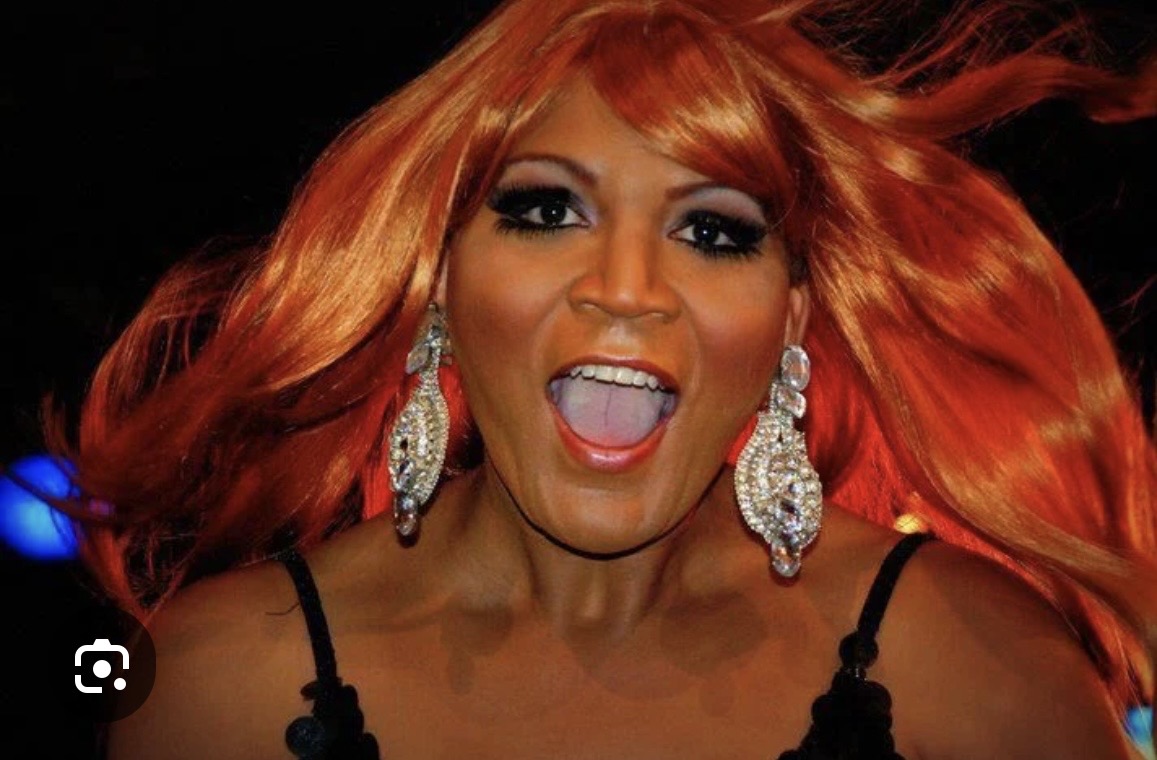 Close-up of a person with vibrant orange hair, bold makeup, and large sparkling earrings smiling joyfully.