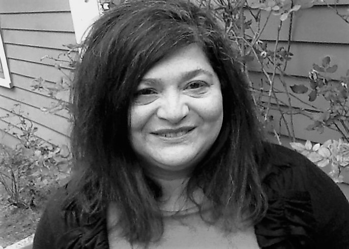 Smiling woman with dark hair standing outdoors near a house with plants and siding in the background