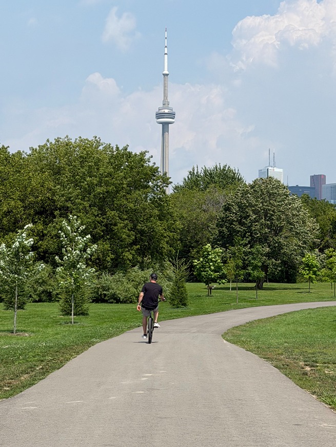 Person cycling on a winding park path with Toronto skyline and CN Tower in the background on a sunny day.