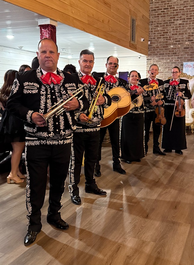 Mariachi band in traditional black charro suits with red bows holding trumpets, guitars, and violins indoors.