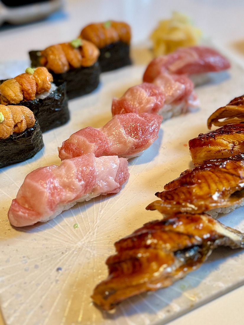 Close-up of assorted sushi including uni, fatty tuna, and eel on a white plate with pickled ginger in the background