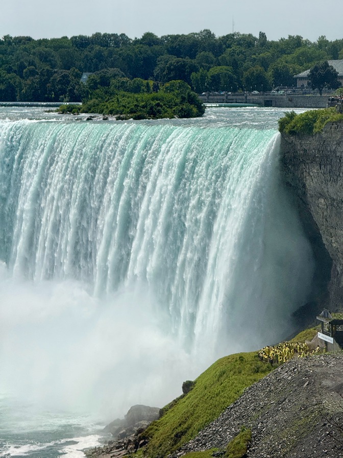 Powerful Niagara Falls cascading over cliff with mist rising and tourists in yellow raincoats on viewing platform.