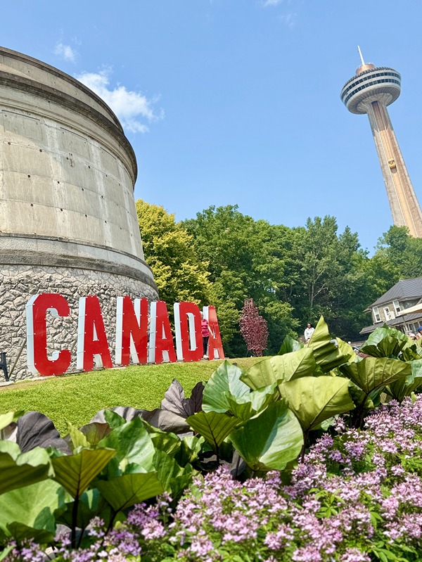 Red Canada sign near a stone tower with green trees and Skylon Tower in Niagara Falls under a blue sky.