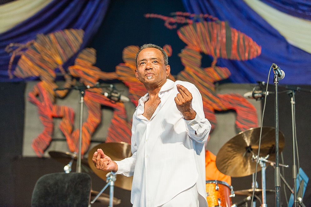 Male jazz singer performing passionately on stage with drum set and colorful backdrop behind him.