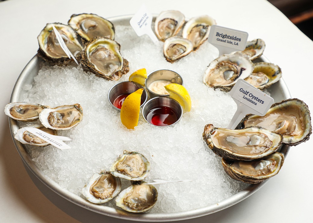 Fresh oysters on ice served with lemon wedges and dipping sauces in a metal tray, labeled by type and origin.