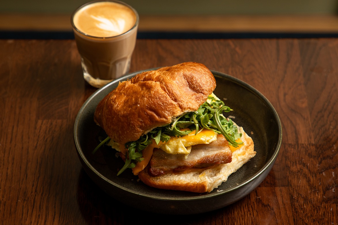 Breakfast croissant sandwich with egg, cheese, greens, and ham served on a black plate with a latte on wooden table.
