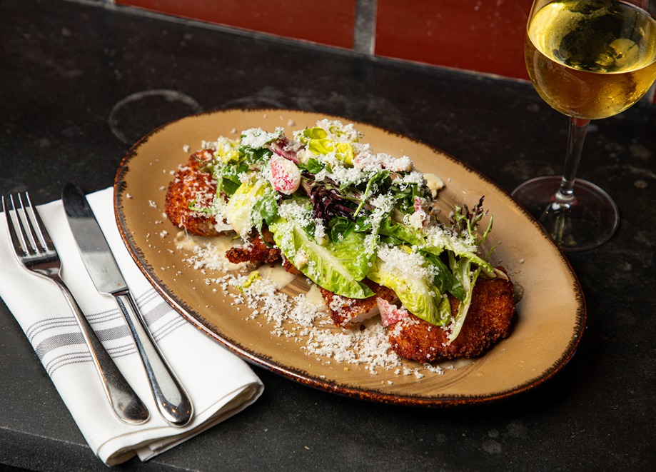 Crispy fried chicken topped with mixed green salad and grated cheese served on a rustic plate with white wine and cutlery.
