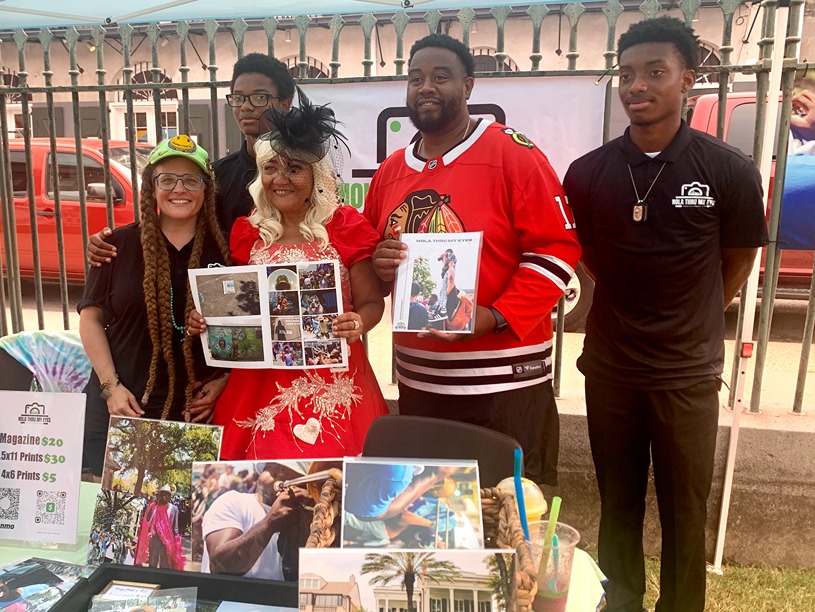 Group of five diverse people displaying photography magazines and prints at outdoor event booth.