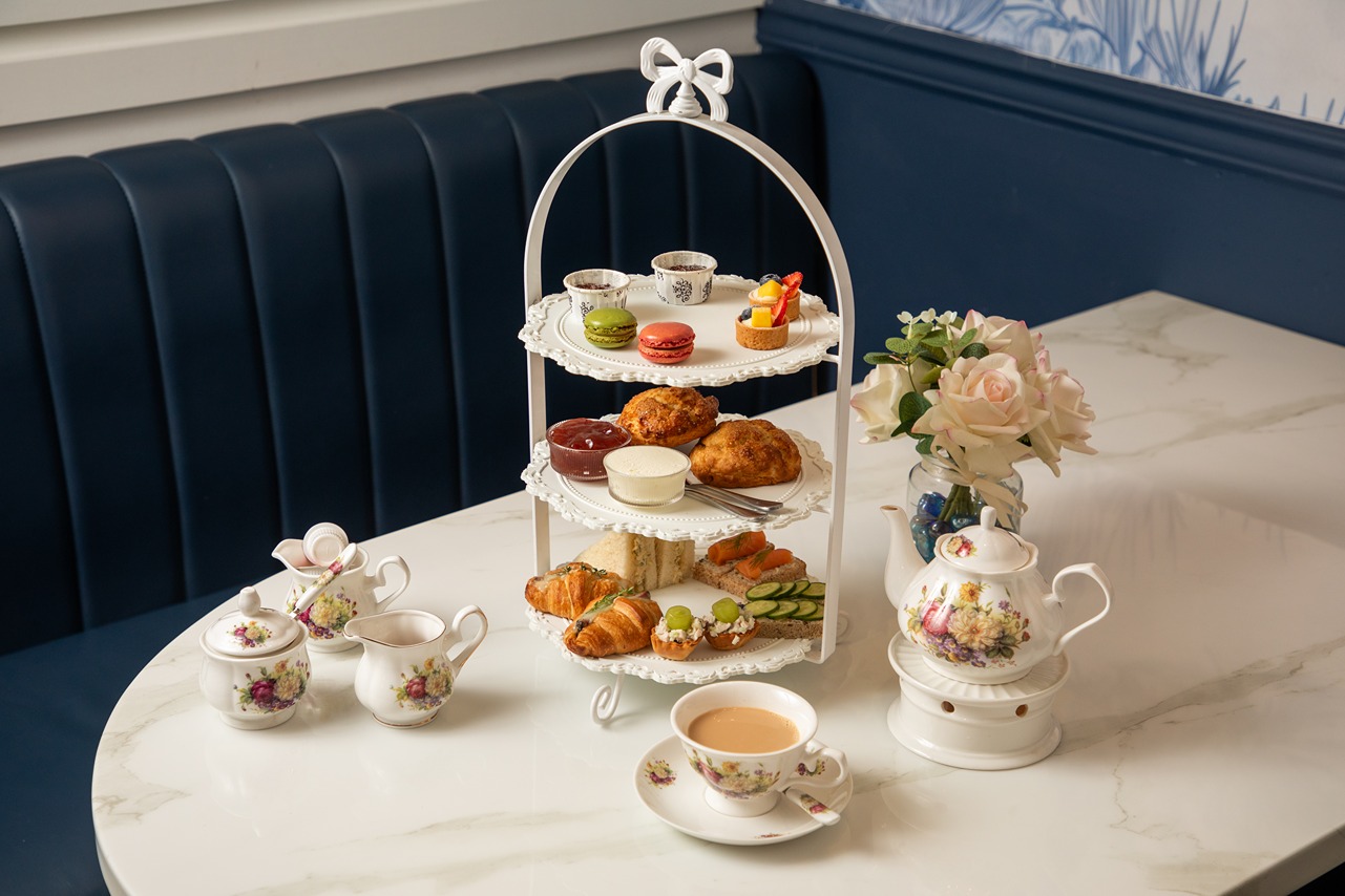 Elegant afternoon tea setup with tiered tray of desserts and sandwiches, fine china, and floral decorations on marble table.