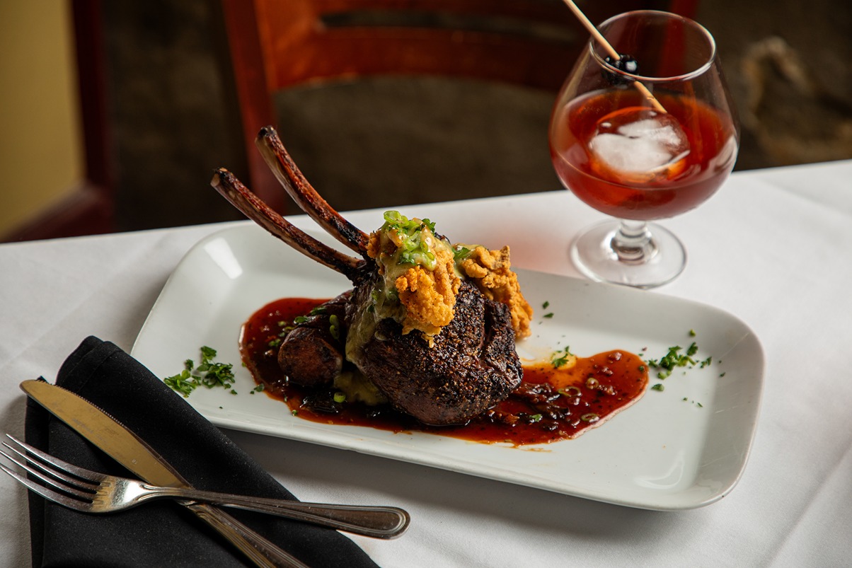 Gourmet rack of lamb with crispy topping and sauce served on a white plate with cocktail on table.