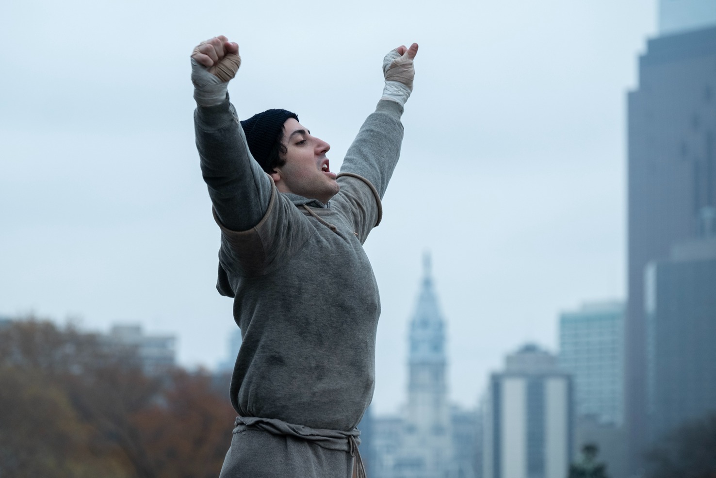 Man in gray workout clothes with raised arms celebrates victory outdoors with cityscape in the background.