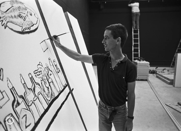 Young male artist painting a large black and white mural of bottles and fish on a wall inside an art studio.