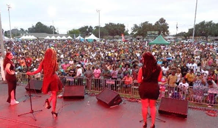 Large outdoor concert with three female performers on stage facing a diverse crowd under cloudy sky.