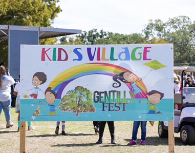 Colorful Kids Village sign with cartoon children playing under a rainbow at Gentilly Fest outdoor event.