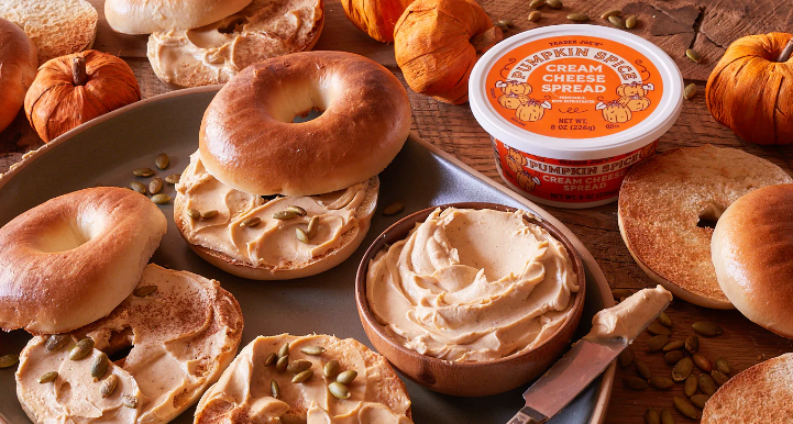Bagels spread with pumpkin spice cream cheese and pumpkin seeds with a container of pumpkin spice cream cheese on a wooden table.
