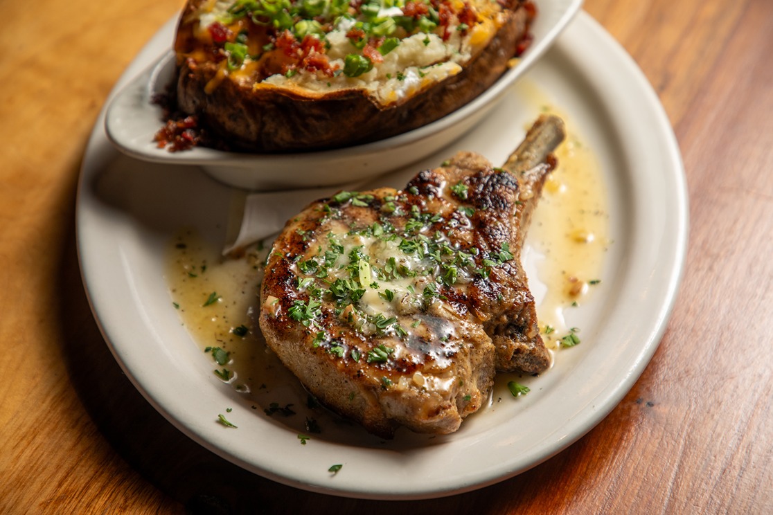 Grilled pork chop with herb sauce and loaded baked potato topped with cheese, bacon, and green onions on white plate.