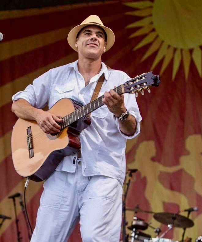 Musician wearing white and a hat plays acoustic guitar onstage with sun-themed backdrop at outdoor concert.