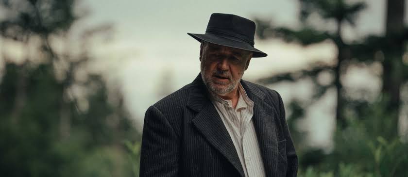 Older man wearing a black hat and striped jacket stands outdoors in a forested area with blurred trees behind him