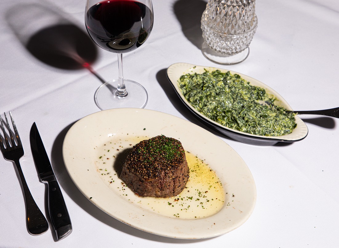 Grilled steak with herbs on white plate, creamy spinach side dish, glass of red wine on white tablecloth.