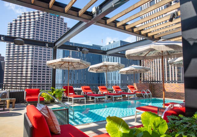Urban rooftop pool area with red lounge chairs, parasols, and modern city buildings in the background