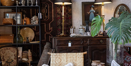 Cozy vintage interior with wooden furniture, woven chairs, brass lamps, decorative plants, and patterned rugs.