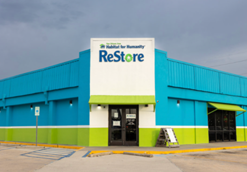 Bright blue and green Habitat for Humanity ReStore building under cloudy sky in an empty parking lot.