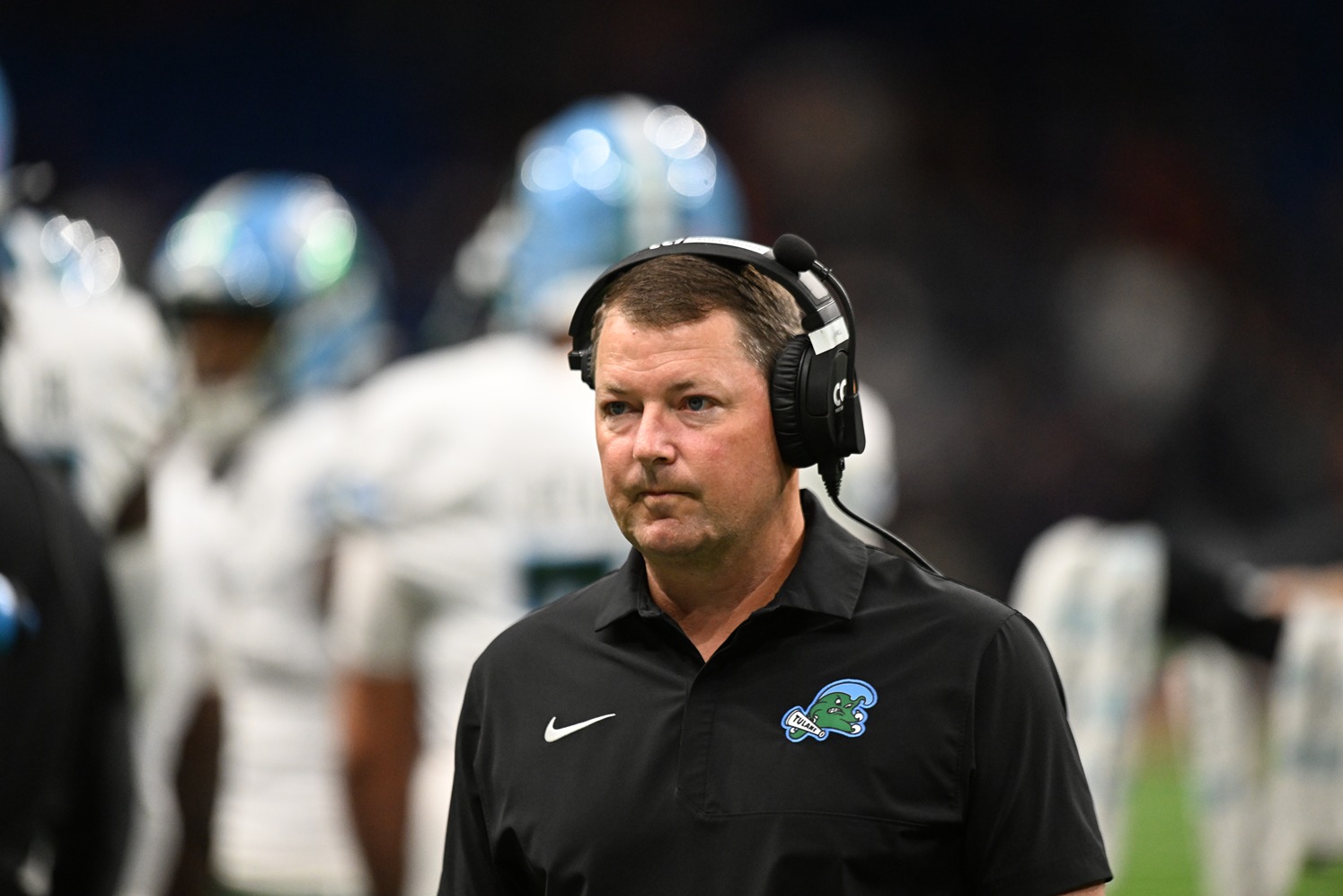 Football coach wearing headset and black polo watches game with blurred players in white uniforms behind him.