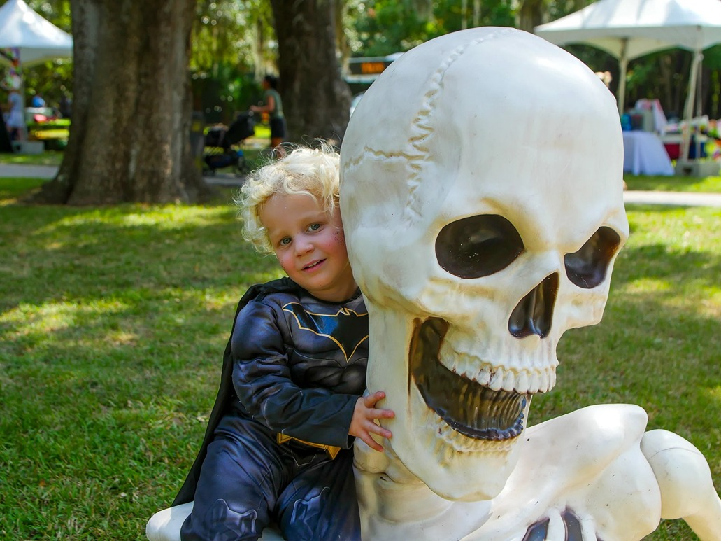 Child dressed as Batman hugging a large smiling skeleton figure outdoors on grass during daytime