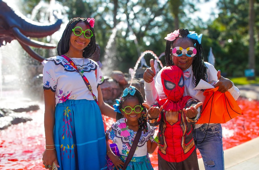 Group of children in colorful costumes and glasses posing happily by a red fountain outdoors on a sunny day