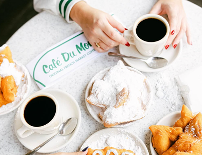 Two cups of black coffee and plates of powdered beignets at Café Du Monde with hands holding a cup.
