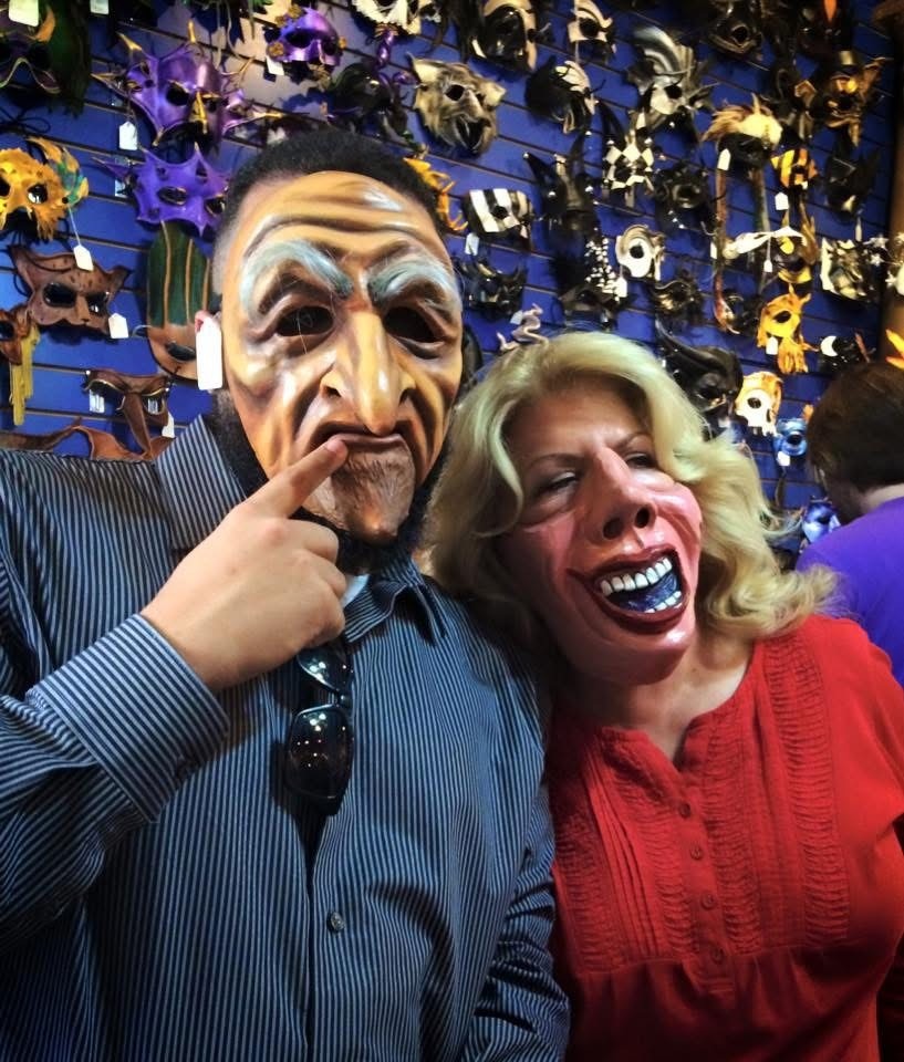 Two people wearing exaggerated, creepy masks in front of a wall covered with various colorful masks.