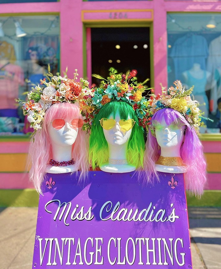 Three mannequins with colorful wigs, floral crowns, and sunglasses displayed outside a vintage clothing store.