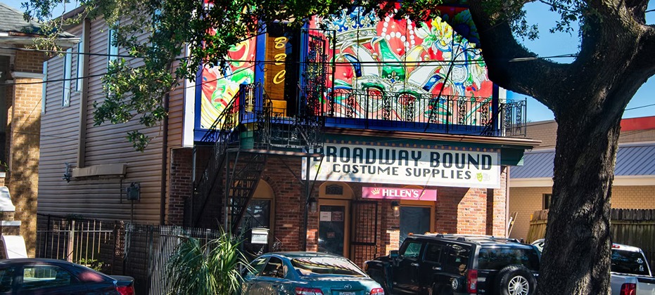 Broadway Bound Costume Supplies storefront with colorful mural, fire escape, parked cars, and tree in urban setting.