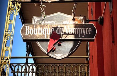 Hanging wooden sign reading Boutique du Vampyre with decorative metal bracket and red wall background.